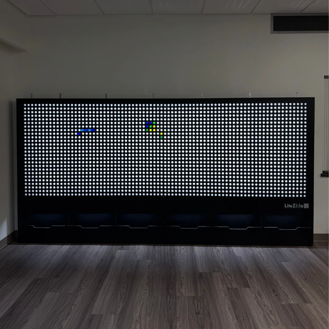 A large backlit wall of small white lights with a few colored pegs pushed into it, standing in an empty room with a wood floor.